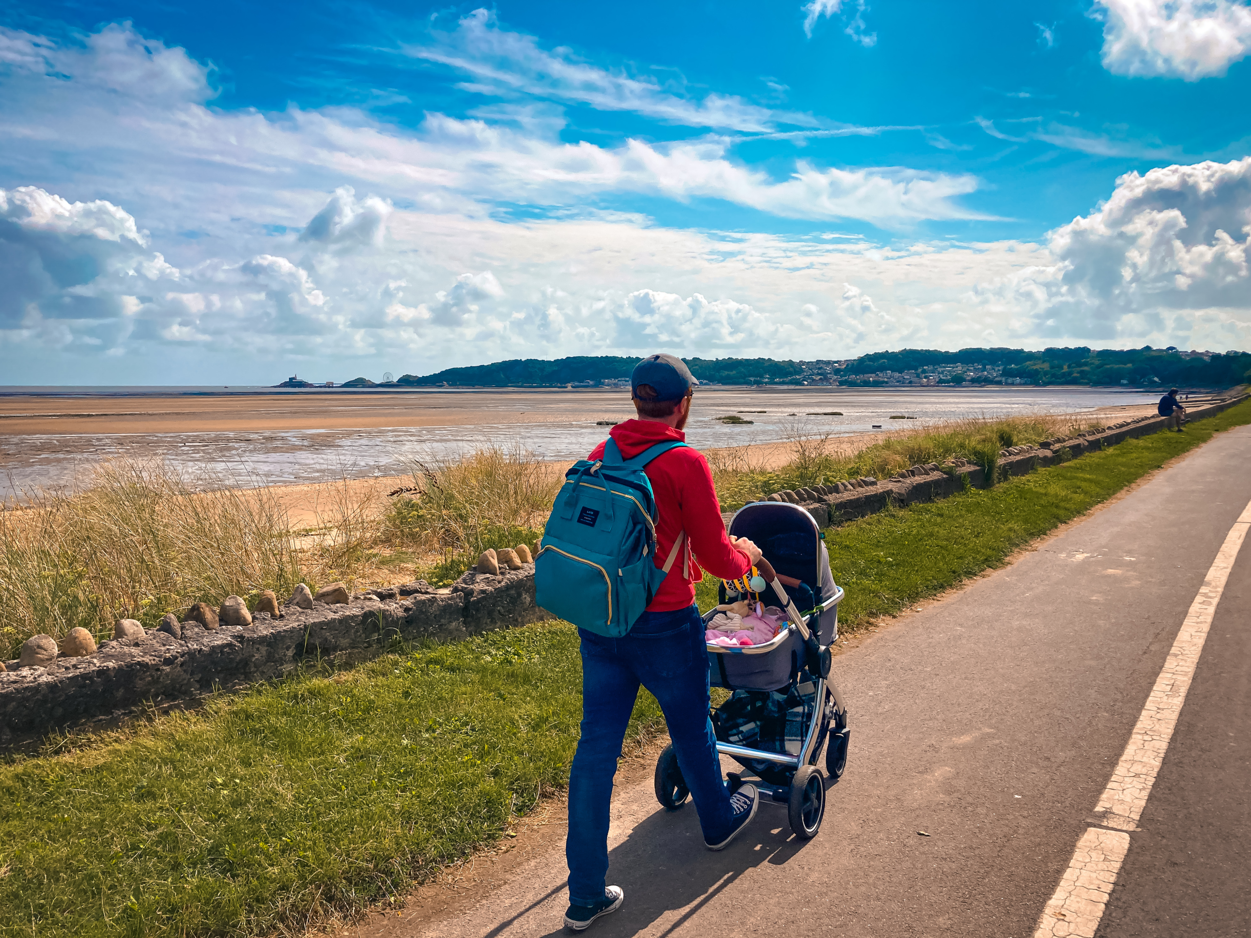 A man pushing a pram along a footpath, with the sea beside him, and view across to Mumbles Pier