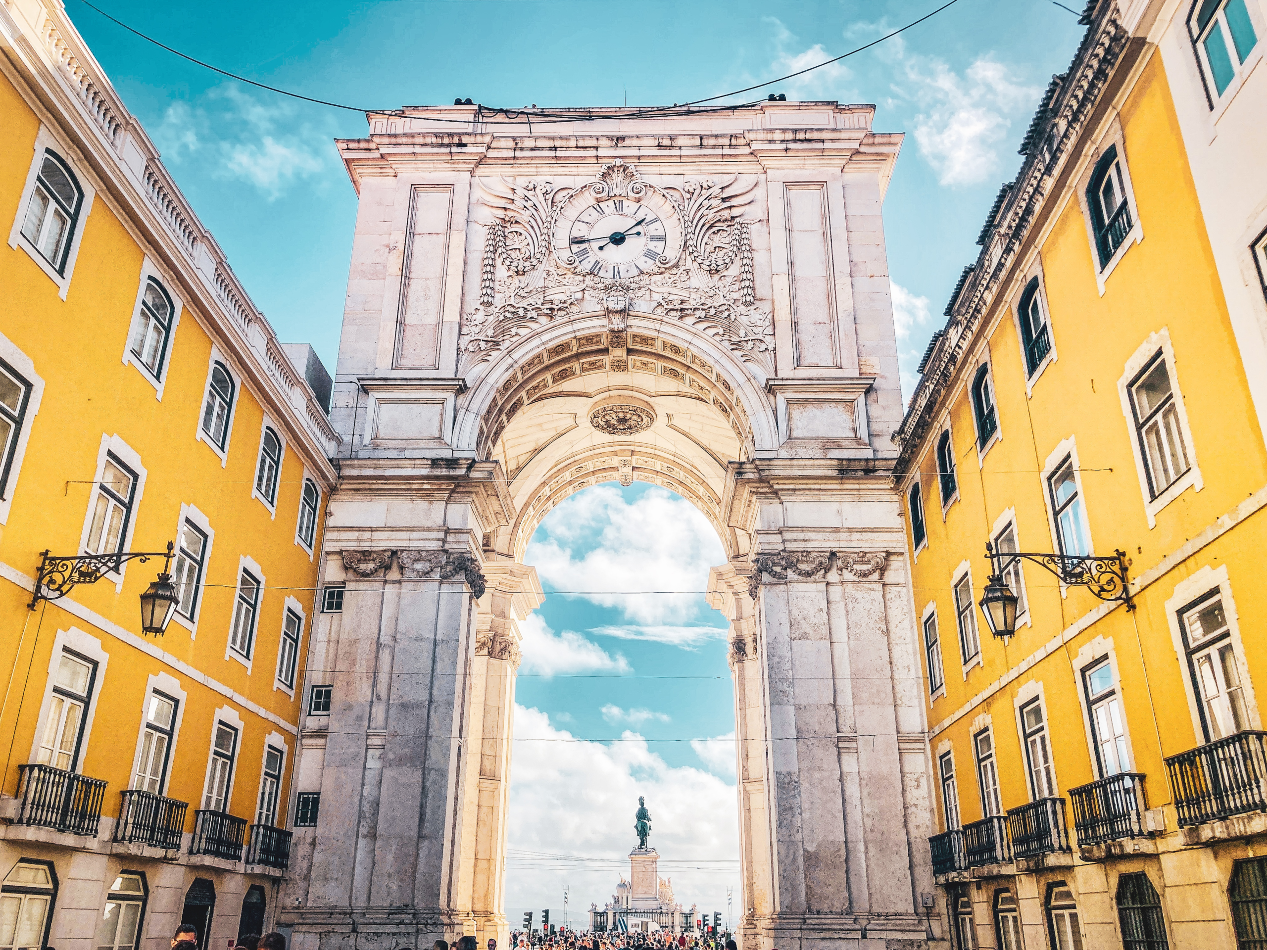 An impressive stone archway with a clock in the centre, lined either side with bright yellow buildings, in Lisbon, Portugal
