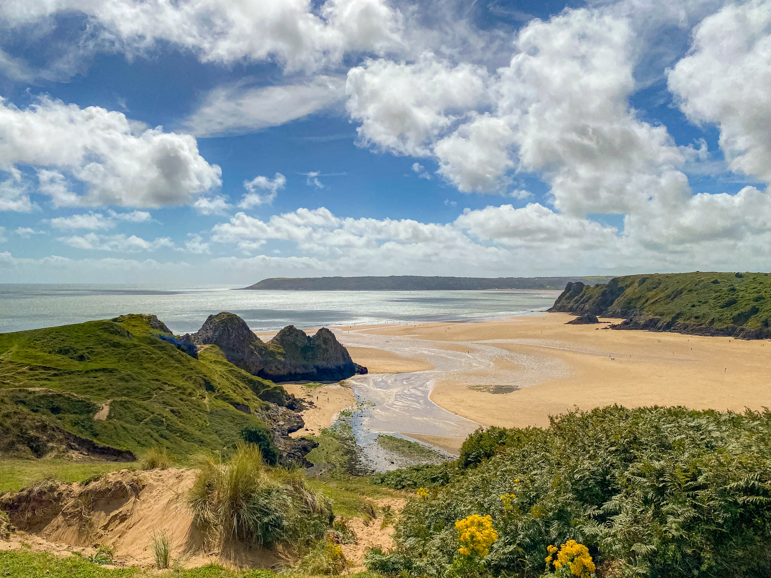 Three cliffs jutting out into the sand, with the sea in the background, surrounded by greenery and hills